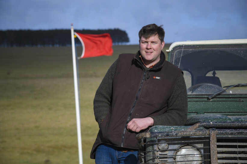 Male farmer leaning on Land Rover with red flag of military firing range in background