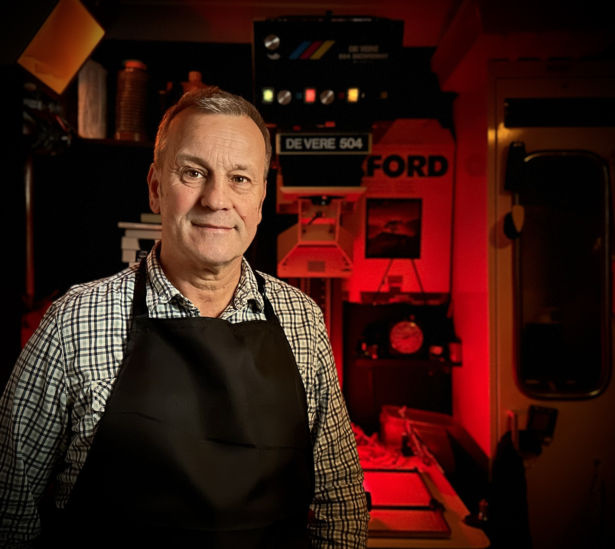 Man wearing black apron in photographic darkroom with Deere 504 enlarger in background